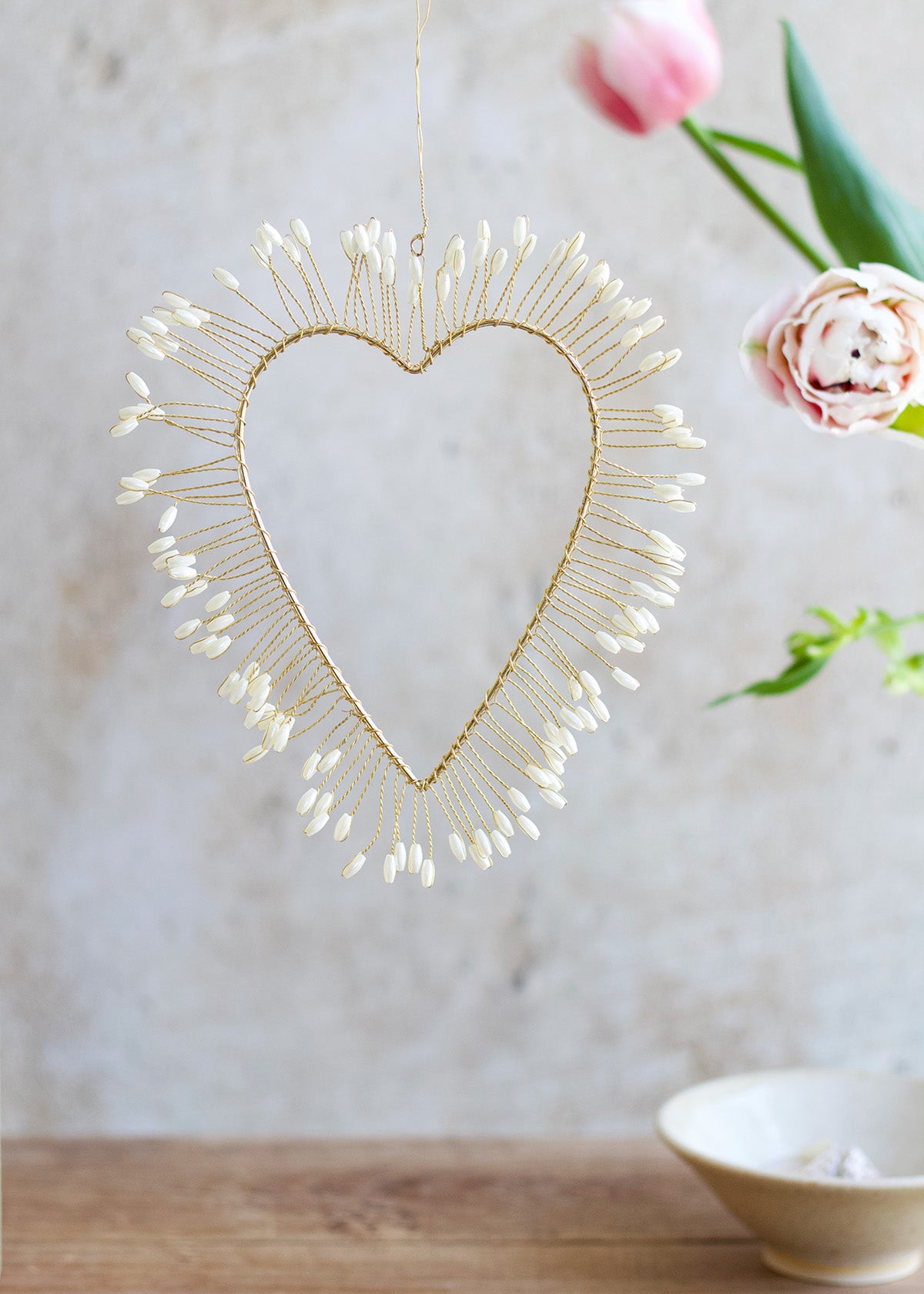 A gold-colored metal heart-shaped hanging ornament with a white pearl-like beaded halo around it, displayed against a neutral background with floral elements and a bowl.