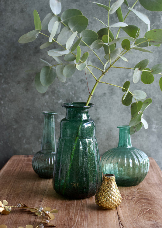 Green glass vases with eucalyptus on a wooden surface against a gray wall