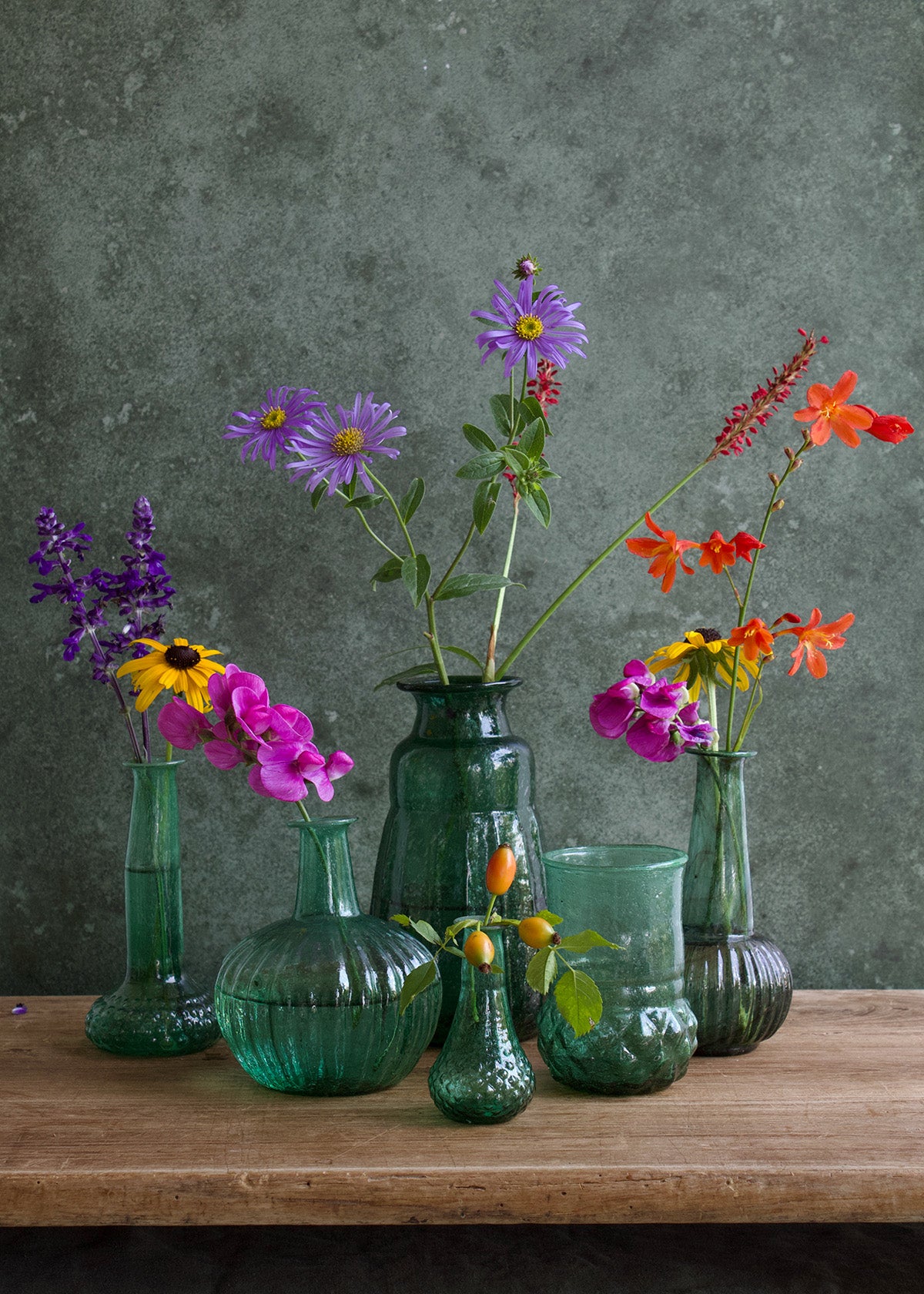 Collection of green glass vases with colorful flowers on a wooden surface against a gray background