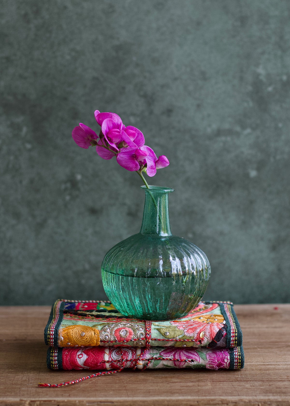 Green textured vase with pink flowers on a colorful fabric against a dark green background