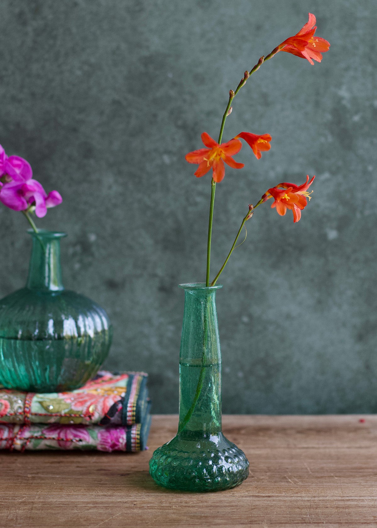 Green vase with orange flowers on a wooden surface against a gray background
