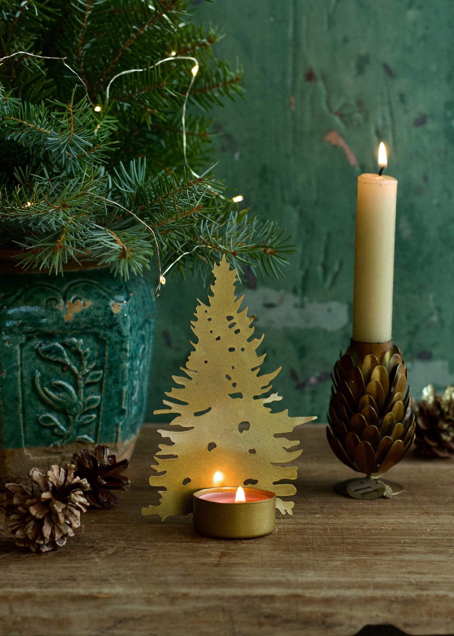 A styled holiday tabletop scene with a gold metal Christmas tree-shaped candle holder holding a small lit tealight candle, next to a tall cream pillar candle in a brown textured holder. A green tree branch with warm fairy lights and pinecones appear in the background against a dark green textured wall.