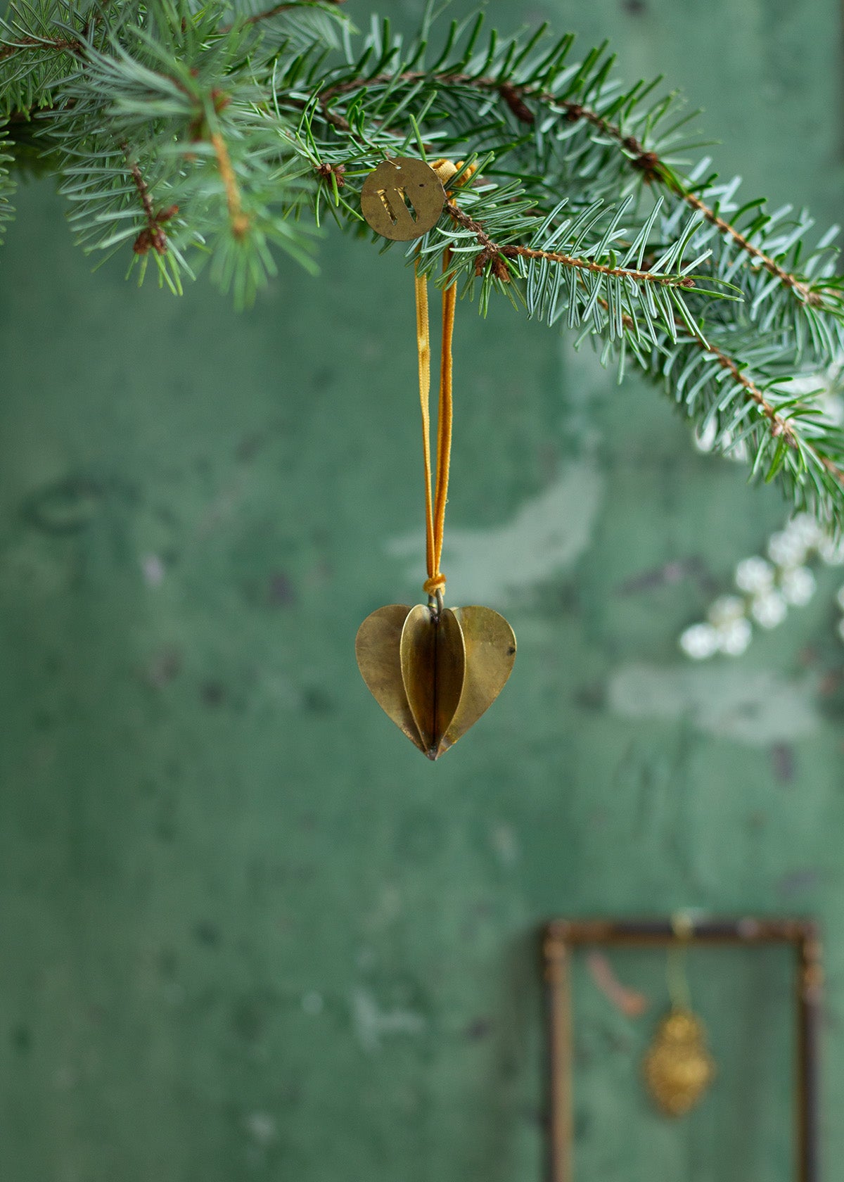 A gold-colored brass hanger shaped like a heart, hanging from a Christmas tree branch against a green background.