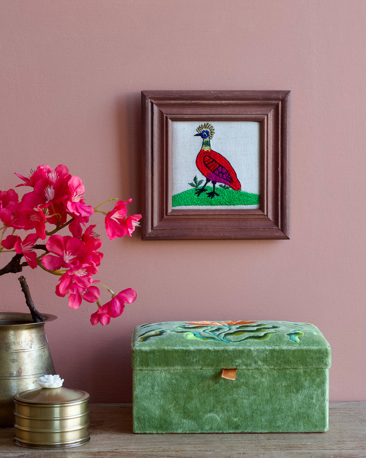 Framed embroidered artwork of a bird on a pink wall with a green box and pink flowers.