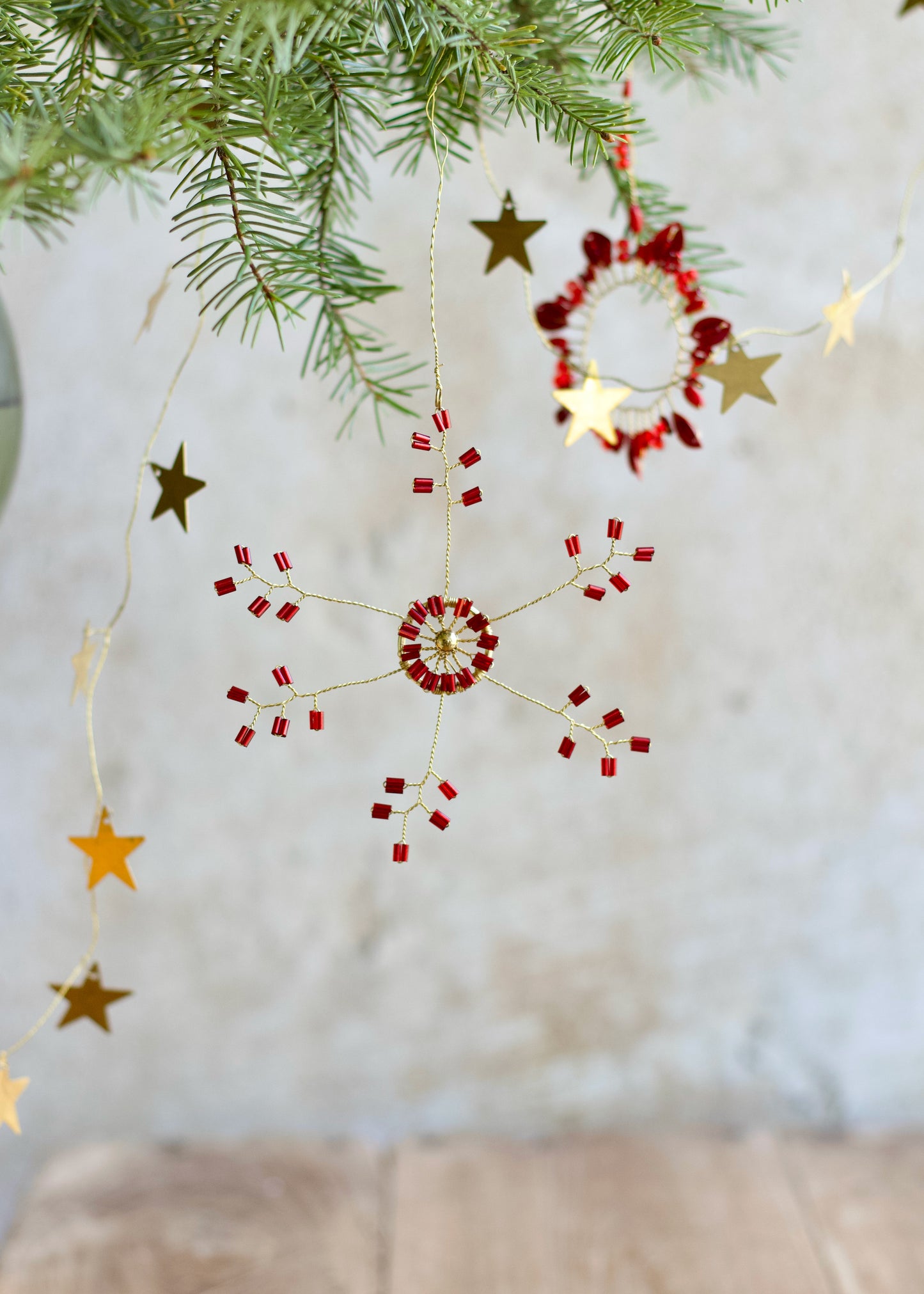 Red and gold beaded snowflake ornament hanging from a pine branch, with brass star garland and a circular ornament in the background.