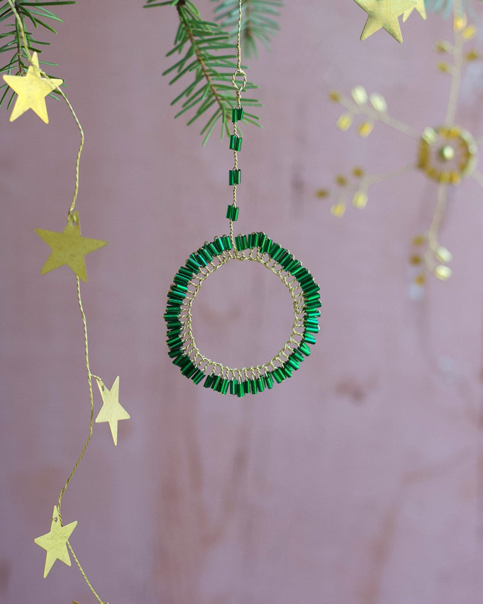Decorative green beaded circle ornament with gold stars hanging from a branch against a soft pink background.