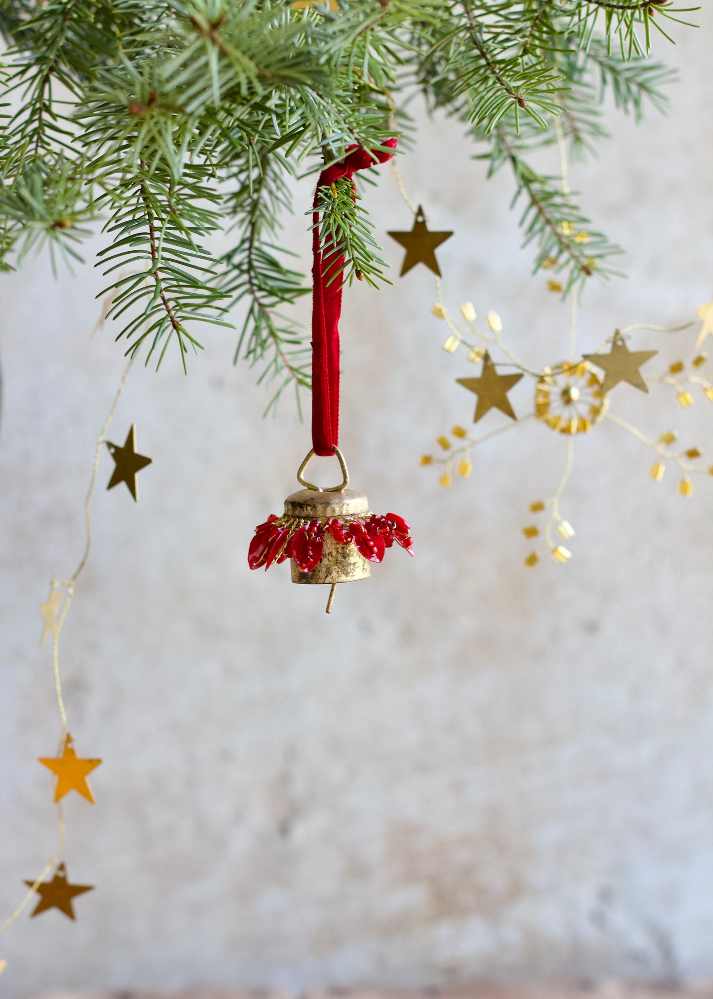 Decorative Christmas bell ornament with red beads hanging on a branch with gold stars and lights.