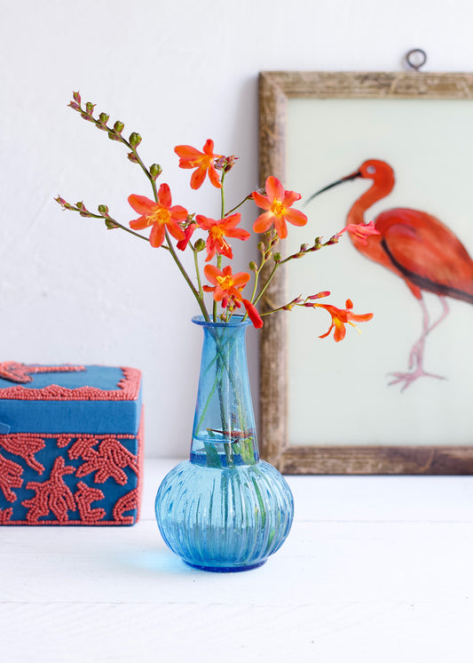 Blue vase with orange flowers on a white surface next to a red box and framed picture of a flamingo.