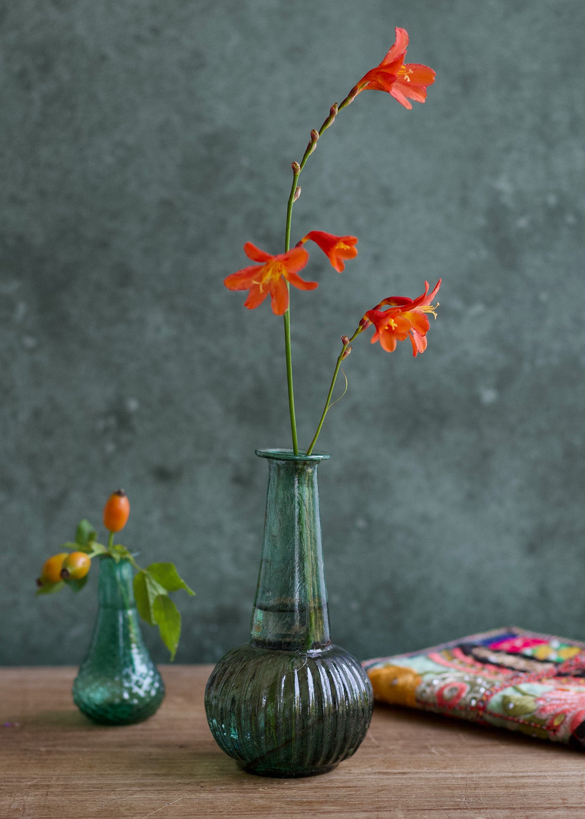 Green vase with orange flowers on a wooden surface against a gray background