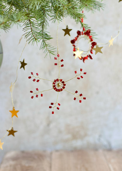 Red and gold beaded snowflake ornament hanging from a pine branch, with brass star garland and a circular ornament in the background.