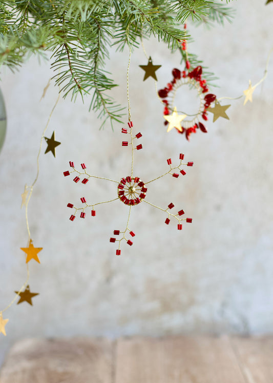Red and gold beaded snowflake ornament hanging from a pine branch, with brass star garland and a circular ornament in the background.