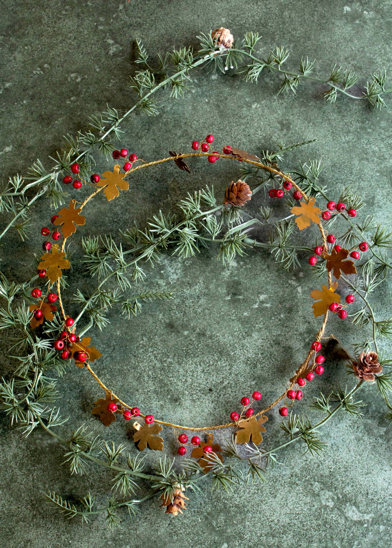 A decorative handmade brass wreath with oak leaves and red berries displayed on a dark textured surface.