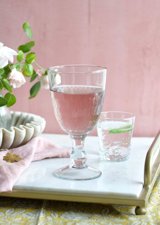 A clear handmade wine glass filled with a pink liquid, placed on a table with a floral patterned napkin and a small vase of flowers in the background.