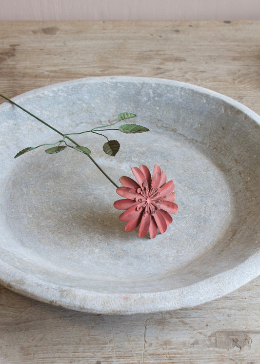 A handmade metal flower with multicolored petals and a green leaf-like design, placed in a beige ceramic vase on a white plate, set against a neutral background.