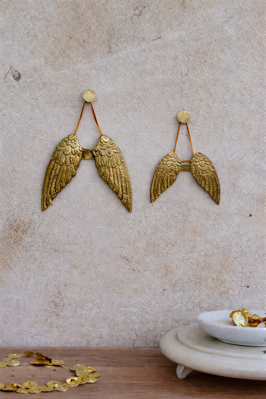 Gold angel wing earrings on a textured beige wall with a small dish and gold leafs at the bottom.