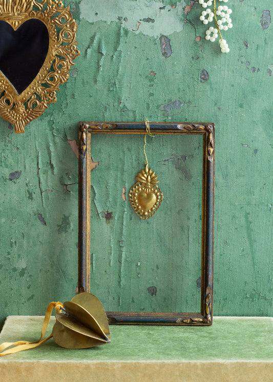 Decorative gold heart pendant hanging in a vintage frame against a textured green wall.