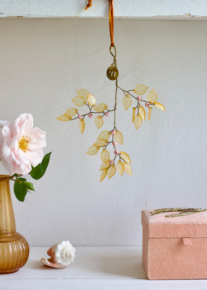 Decorative hanging branch with yellow leaves next to a pink flower in a vase and a pink box on a light surface.