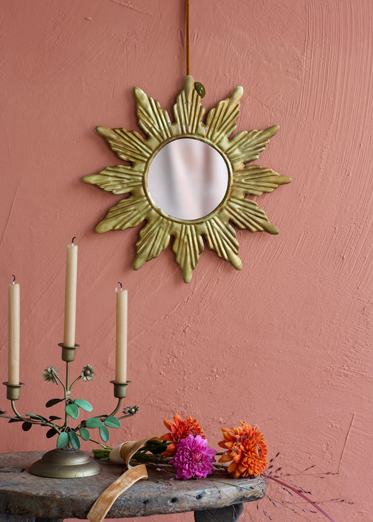 Decorative sun-shaped mirror on a pink wall with candles and flowers on a table.