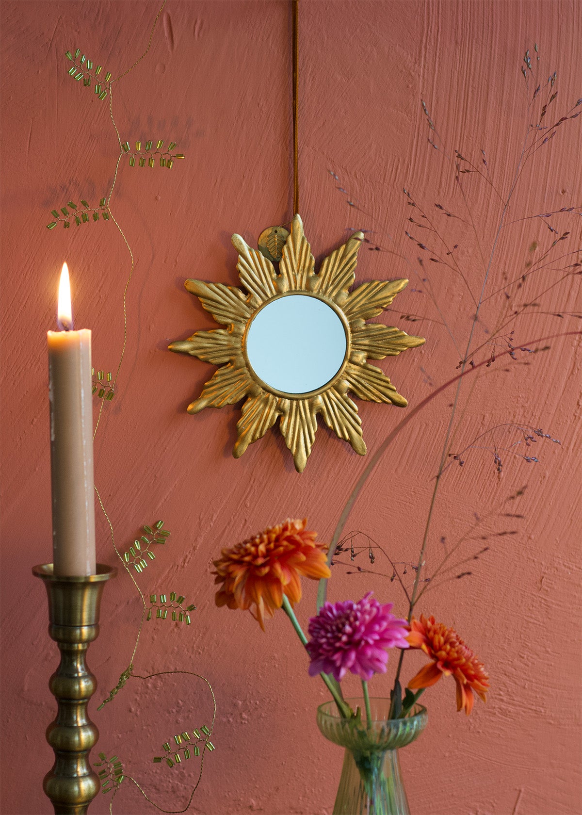 Decorative gold sunburst mirror on a pink wall with a candle and flowers.